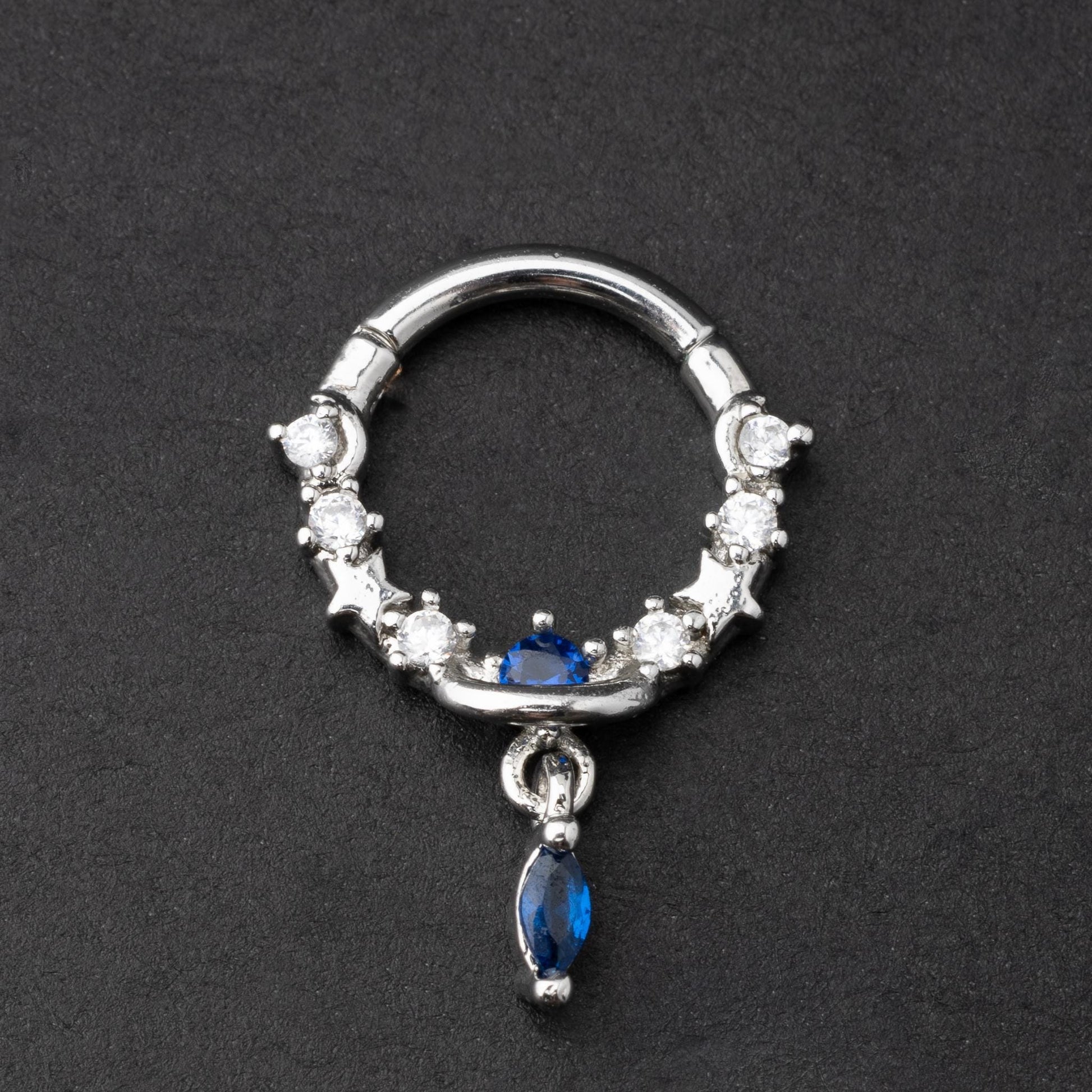 Gold and silver septum clickers adorned with cubic zirconia and a dangling blue gemstone charm, displayed on a dark background.
