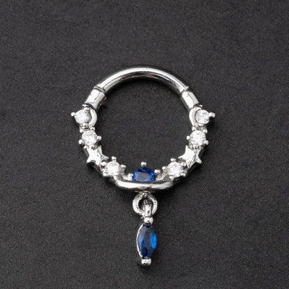Gold and silver septum clickers adorned with cubic zirconia and a dangling blue gemstone charm, displayed on a dark background.