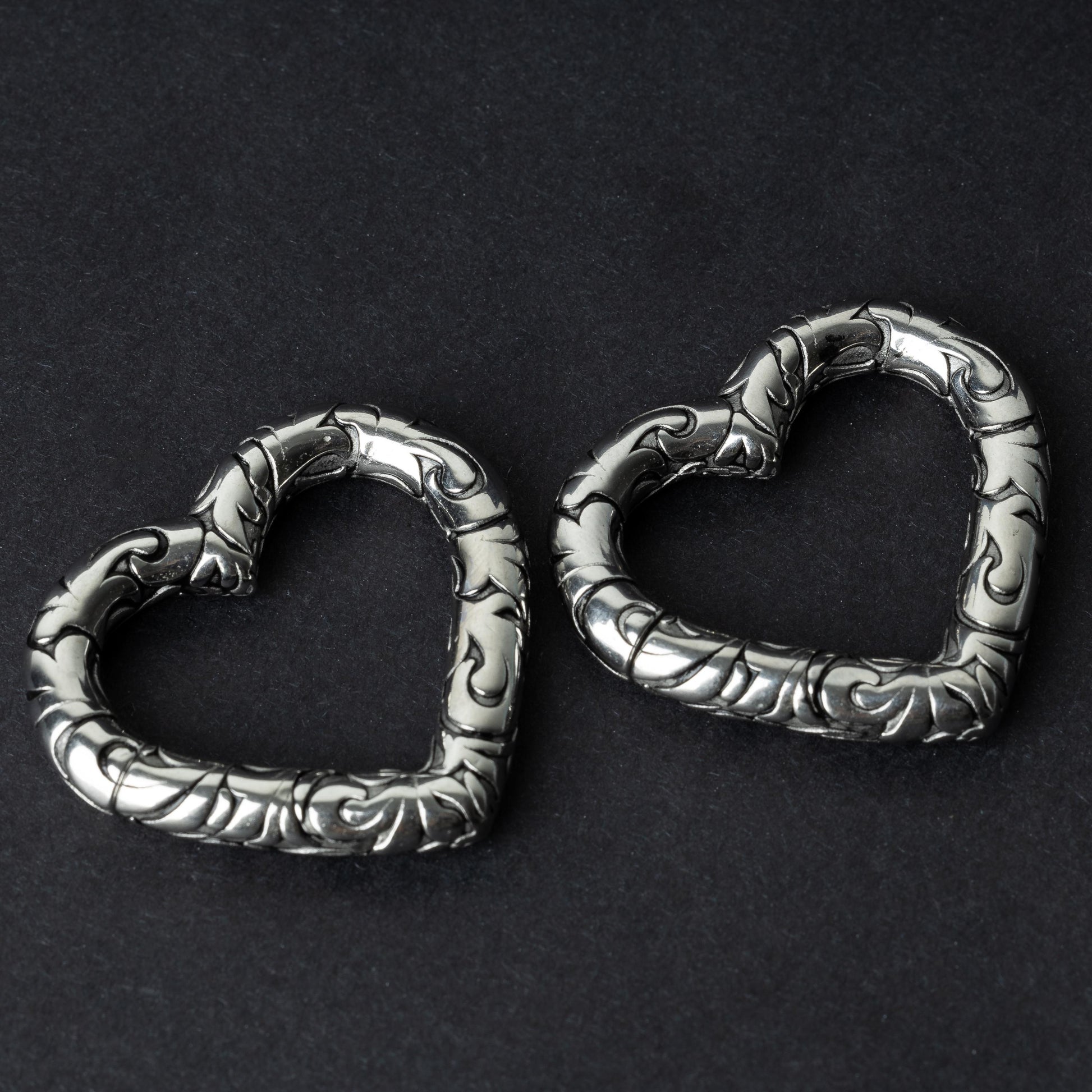 Two heart-shaped ear weights in silver, featuring intricate detailing, displayed on a dark background.

