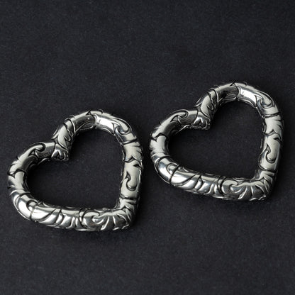 Two heart-shaped ear weights in silver, featuring intricate detailing, displayed on a dark background.

