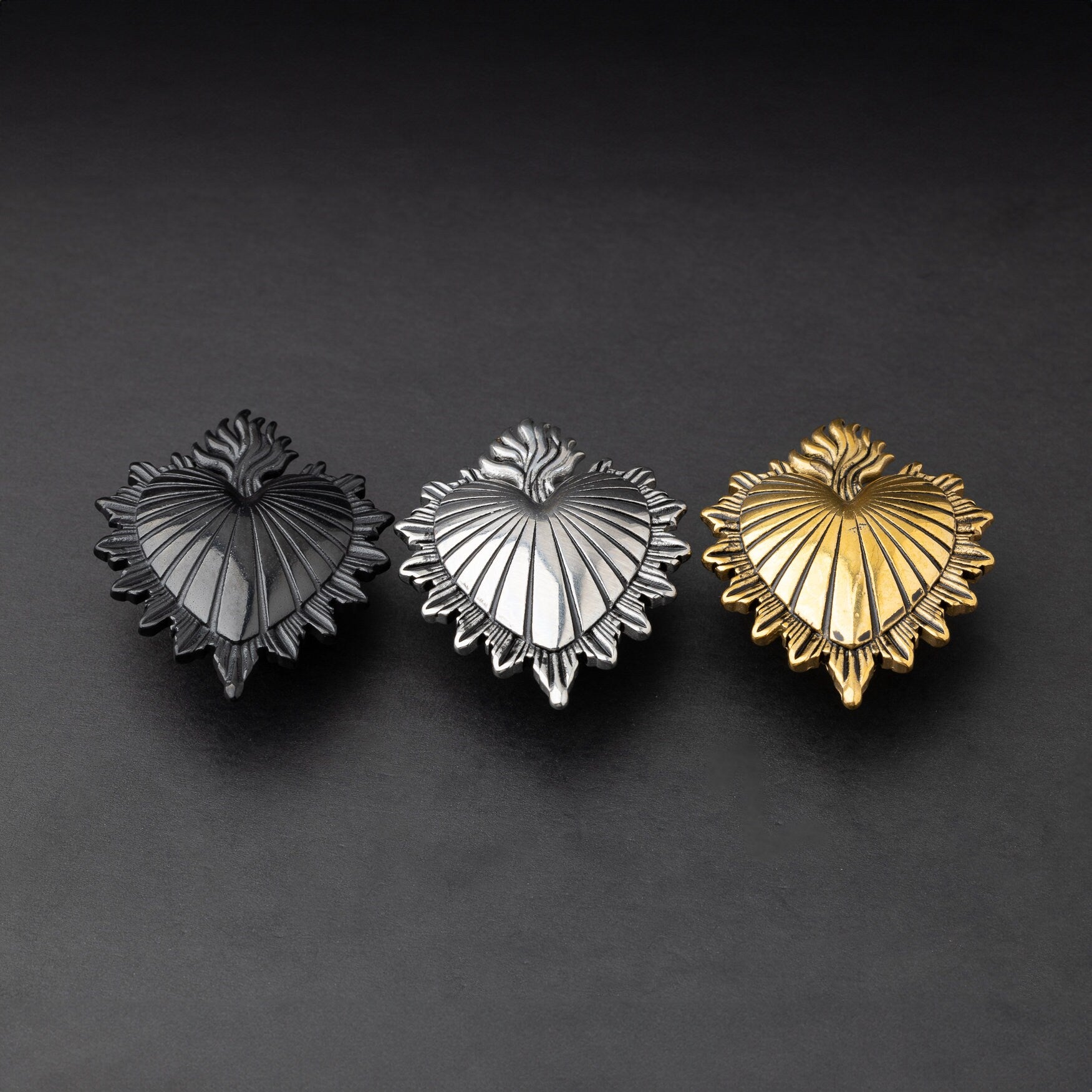 Three Sacred Heart-inspired plugs in black, silver, and gold tones, displayed on a matte dark background, showcasing intricate detailing and a bold design.
