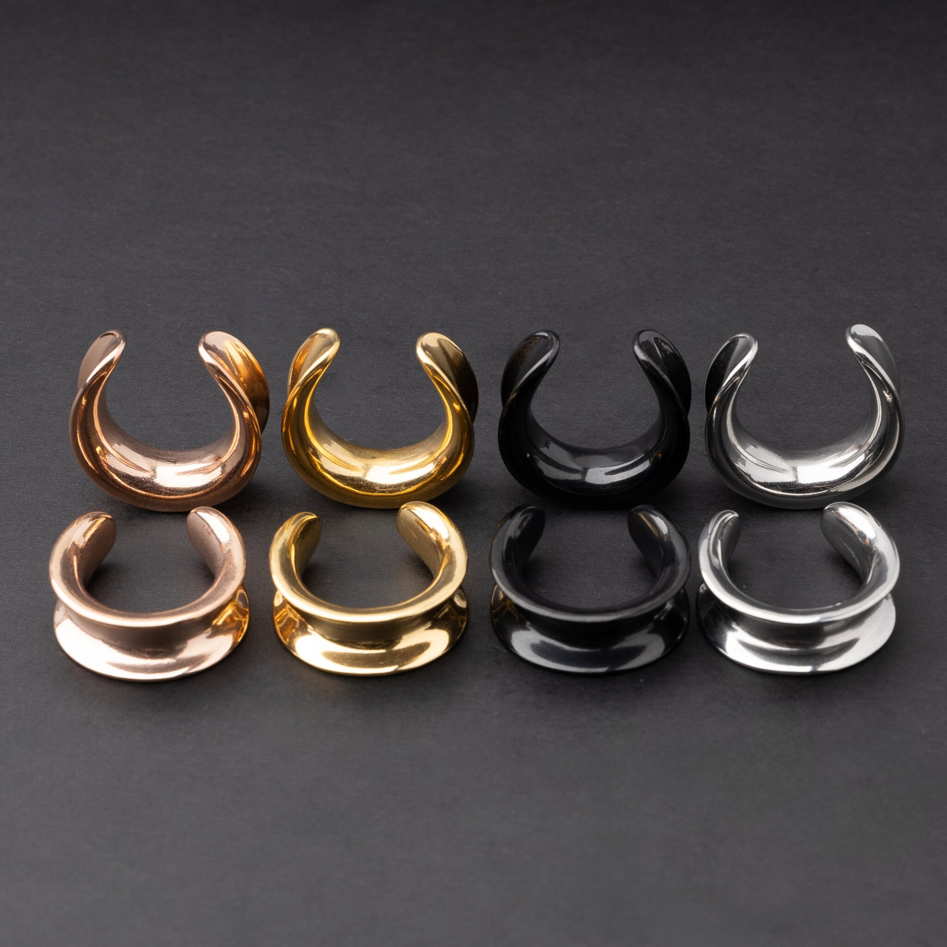 Polished saddle-style tunnels in rose gold, gold, black, and silver finishes, arranged on a matte dark background, showcasing their sleek and minimalist design.
