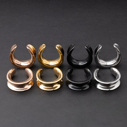 Polished saddle-style tunnels in rose gold, gold, black, and silver finishes, arranged on a matte dark background, showcasing their sleek and minimalist design.