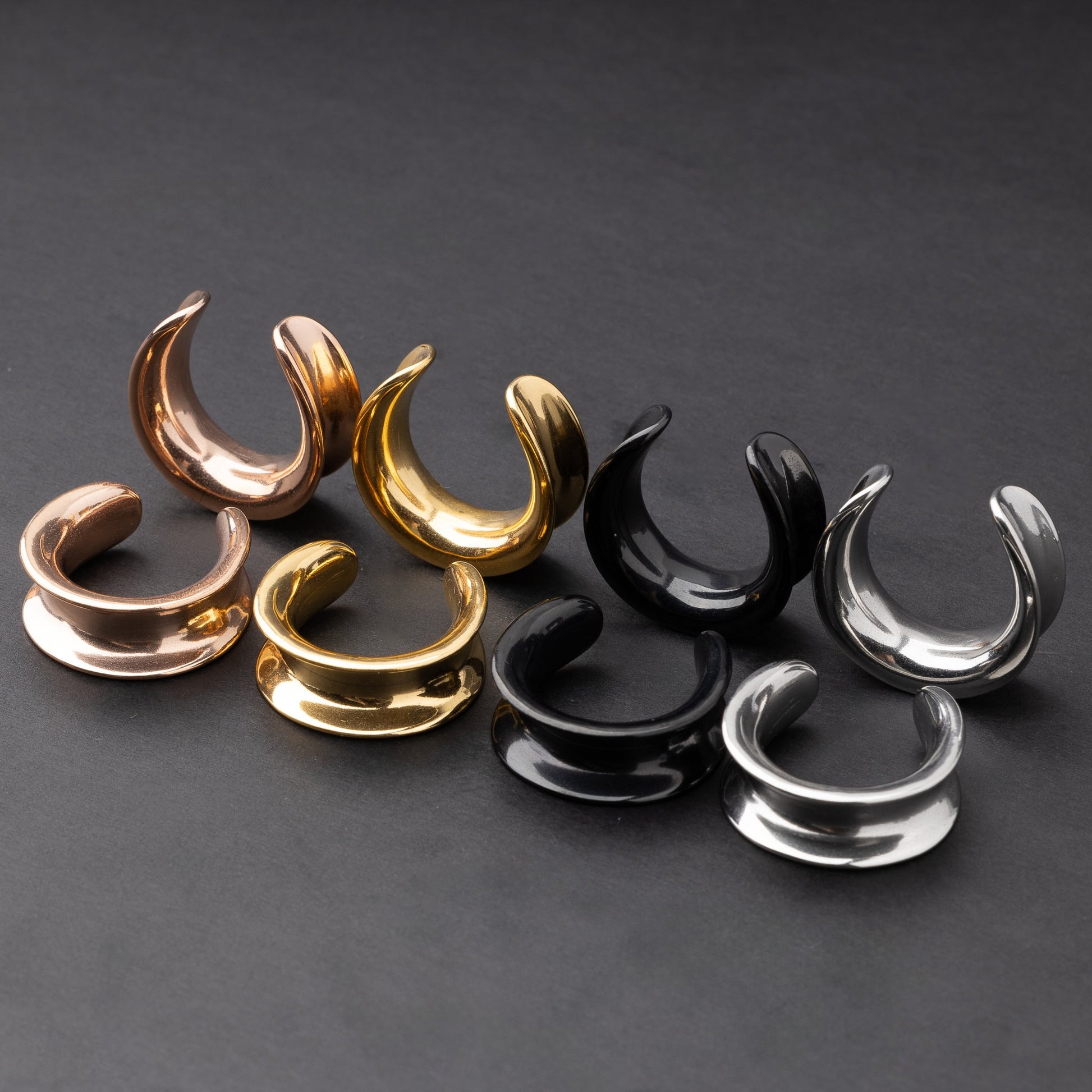 Polished saddle-style tunnels in rose gold, gold, black, and silver finishes, arranged on a matte dark background, showcasing their sleek and minimalist design.