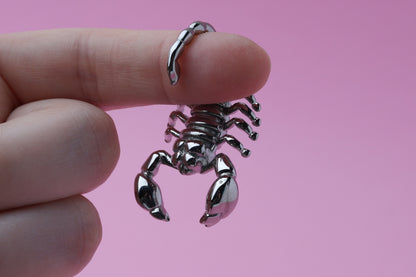 Scorpion-shaped ear hanger in silver and hung from a finger in front of a pink background, highlighting their detailed and edgy design.