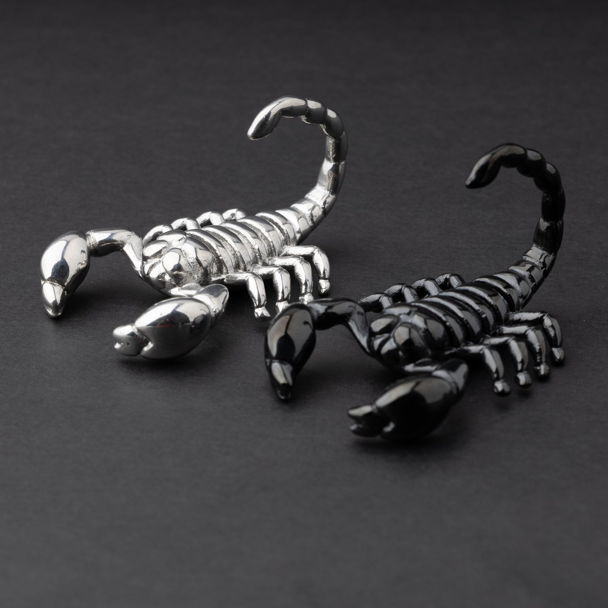 Two scorpion-shaped ear hangers, one in silver and one in black, displayed on a dark matte background, highlighting their detailed and edgy design.