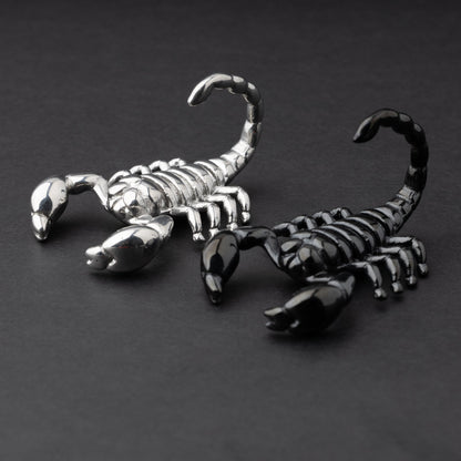 Two scorpion-shaped ear hangers, one in silver and one in black, displayed on a dark matte background, highlighting their detailed and edgy design.