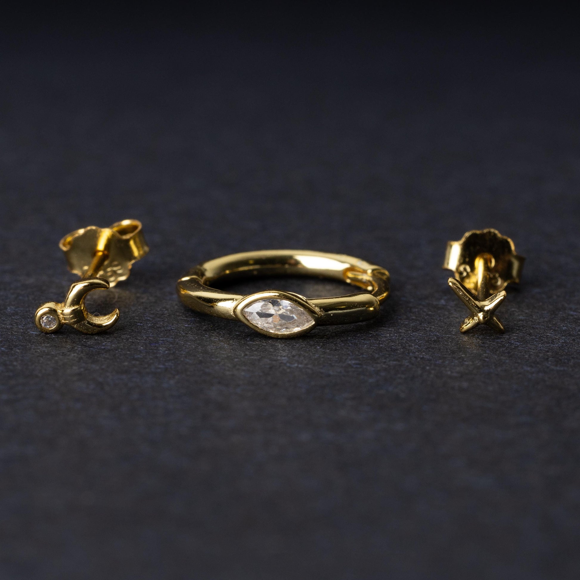 A gold-tone earring set displayed on a dark matte background, featuring a hoop with a marquise-cut crystal, a crescent moon stud with a crystal accent, and a minimalist cross stud.