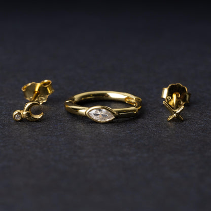 A gold-tone earring set displayed on a dark matte background, featuring a hoop with a marquise-cut crystal, a crescent moon stud with a crystal accent, and a minimalist cross stud.