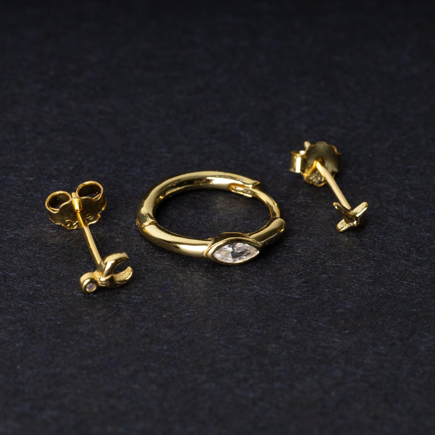 A gold-tone earring set displayed on a dark matte background, featuring a hoop with a marquise-cut crystal, a crescent moon stud with a crystal accent, and a minimalist cross stud.