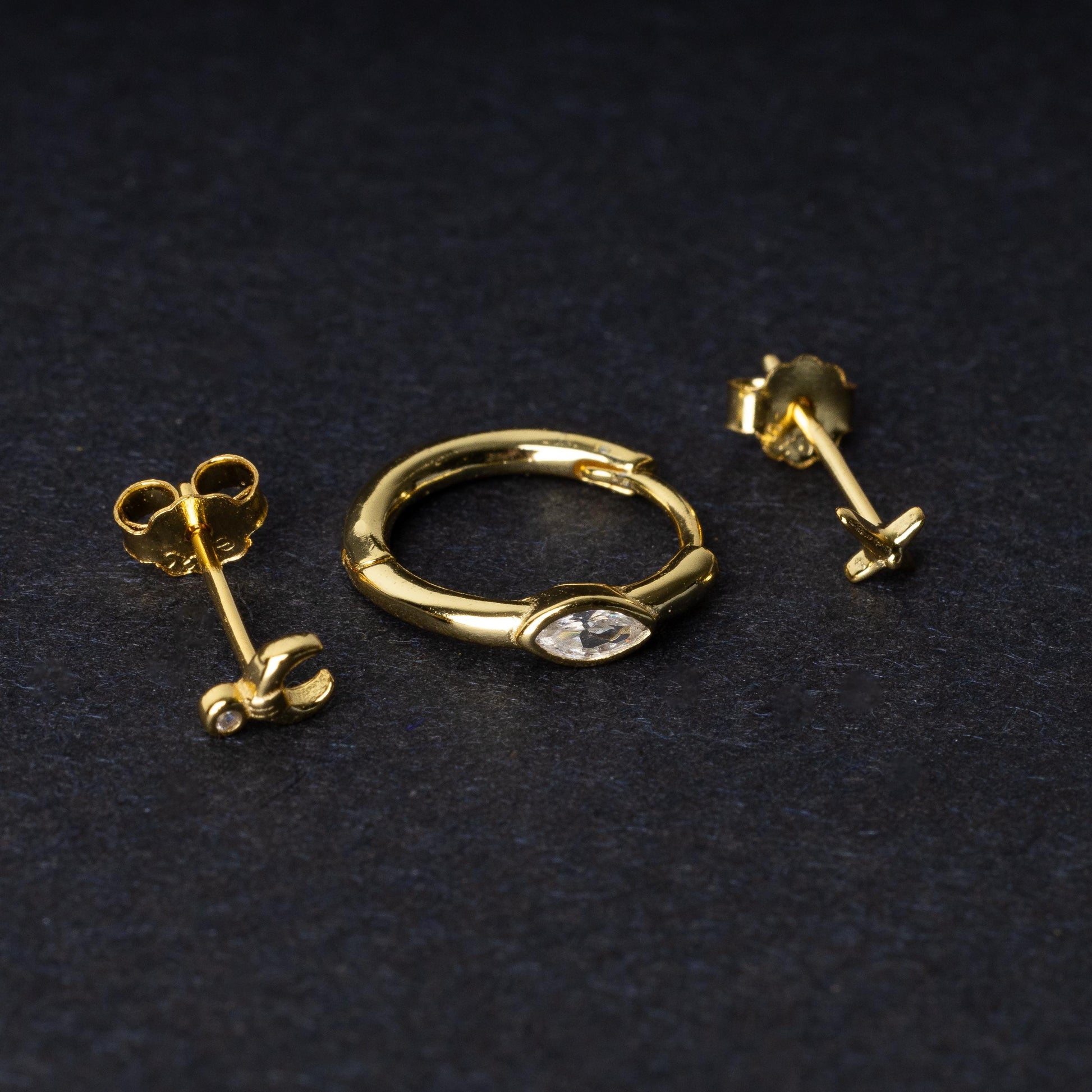 A gold-tone earring set displayed on a dark matte background, featuring a hoop with a marquise-cut crystal, a crescent moon stud with a crystal accent, and a minimalist cross stud.