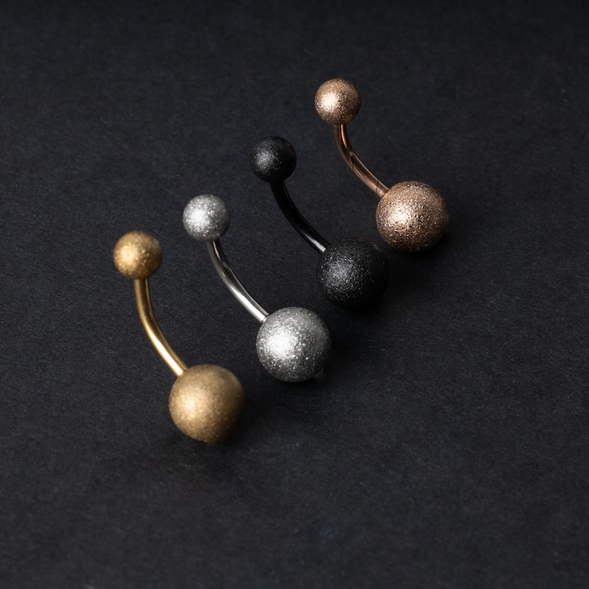 Four curved barbells with shimmering textured ball ends in gold, silver, black, and rose gold finishes, displayed on a dark matte background.