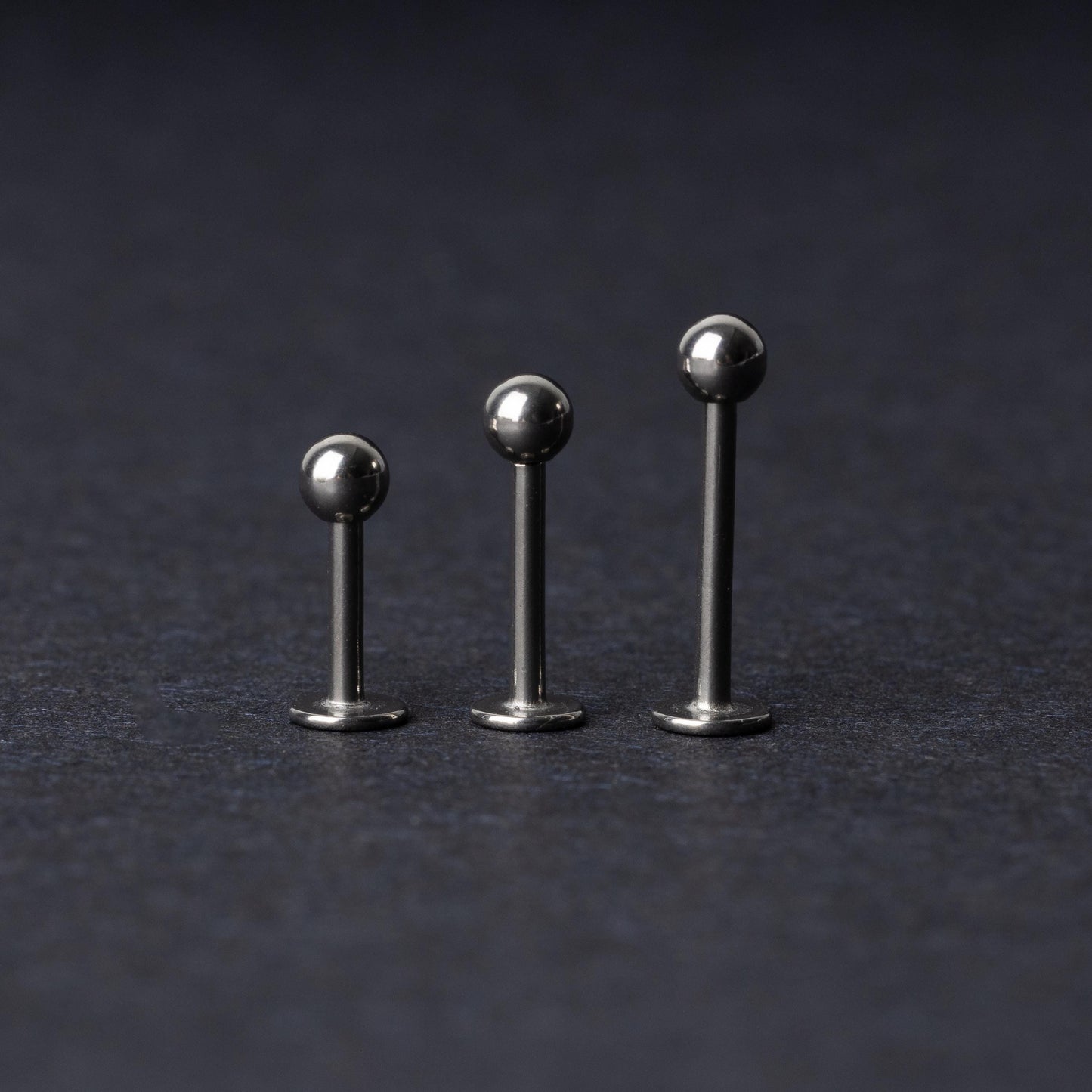 Three silver stainless steel labret studs with flat backs and polished ball ends, displayed on a dark textured background.