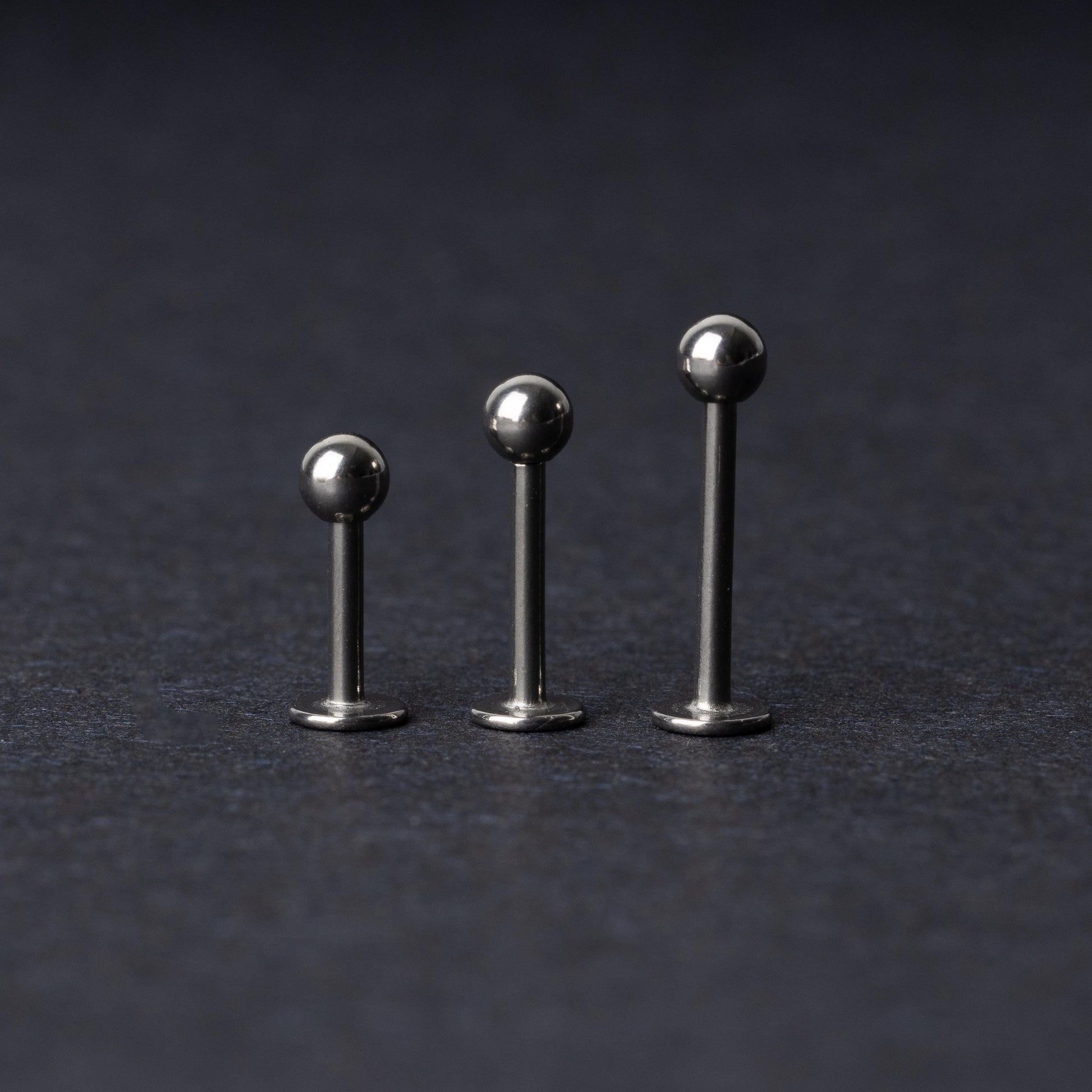 Three silver stainless steel labret studs with flat backs and polished ball ends, displayed on a dark textured background.