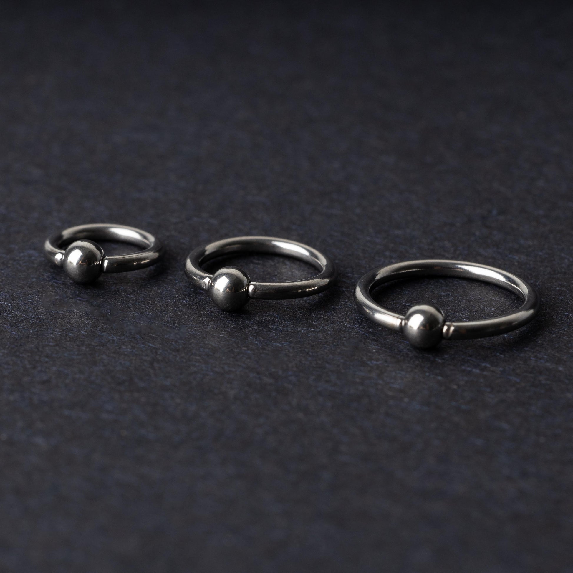 Three polished stainless steel captive bead rings of varying sizes displayed on a dark matte background.
