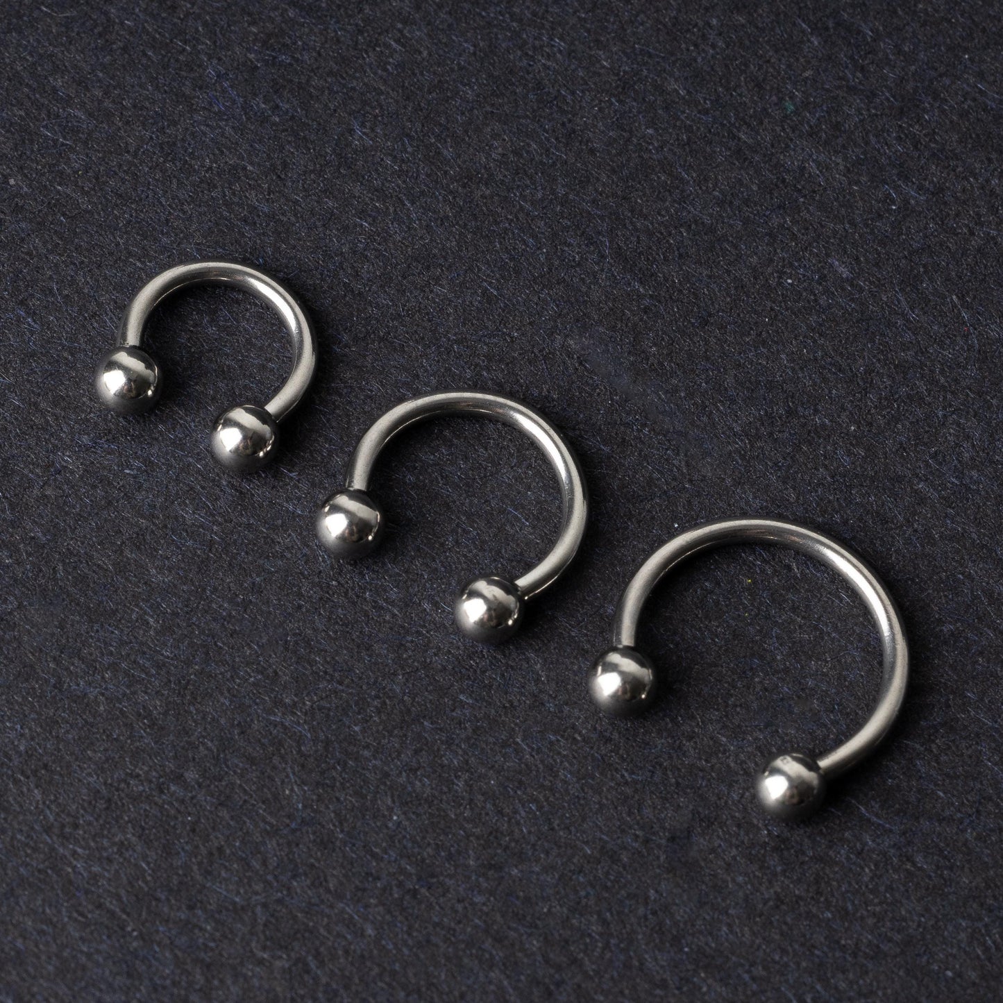 Three silver stainless steel horseshoe hoops with polished ball ends, displayed on a dark textured background.