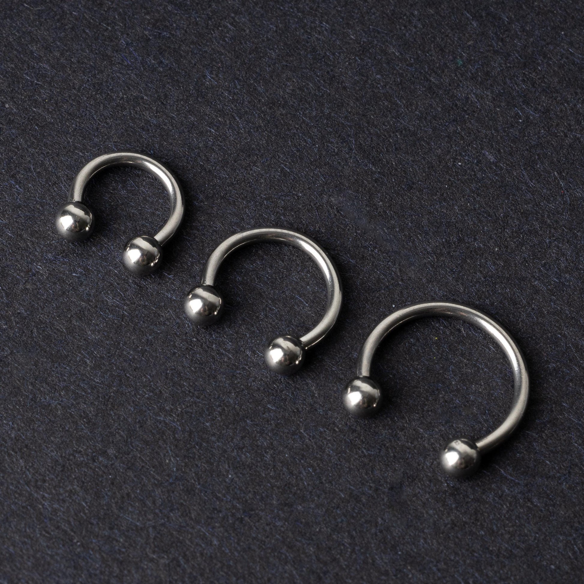 Three silver stainless steel horseshoe hoops with polished ball ends, displayed on a dark textured background.