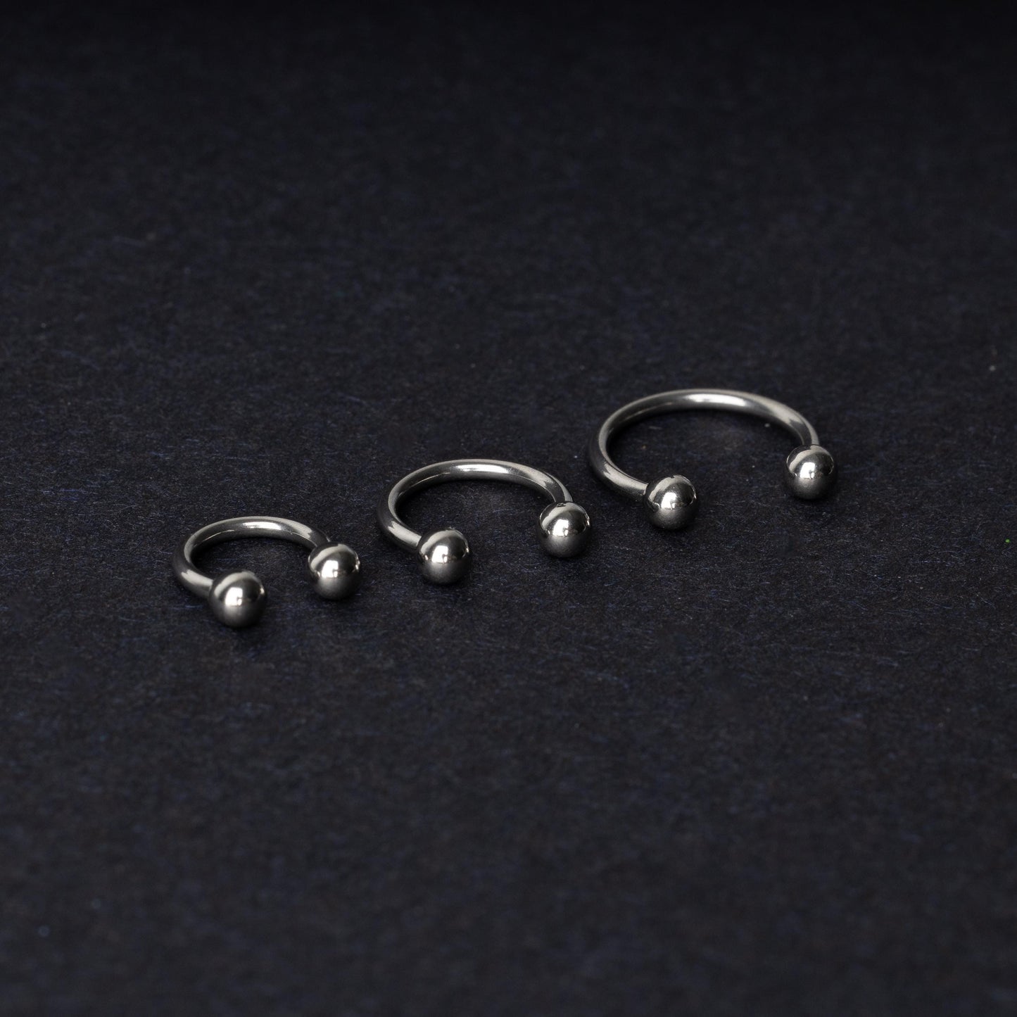 Three silver stainless steel horseshoe hoops with polished ball ends, displayed on a dark textured background.