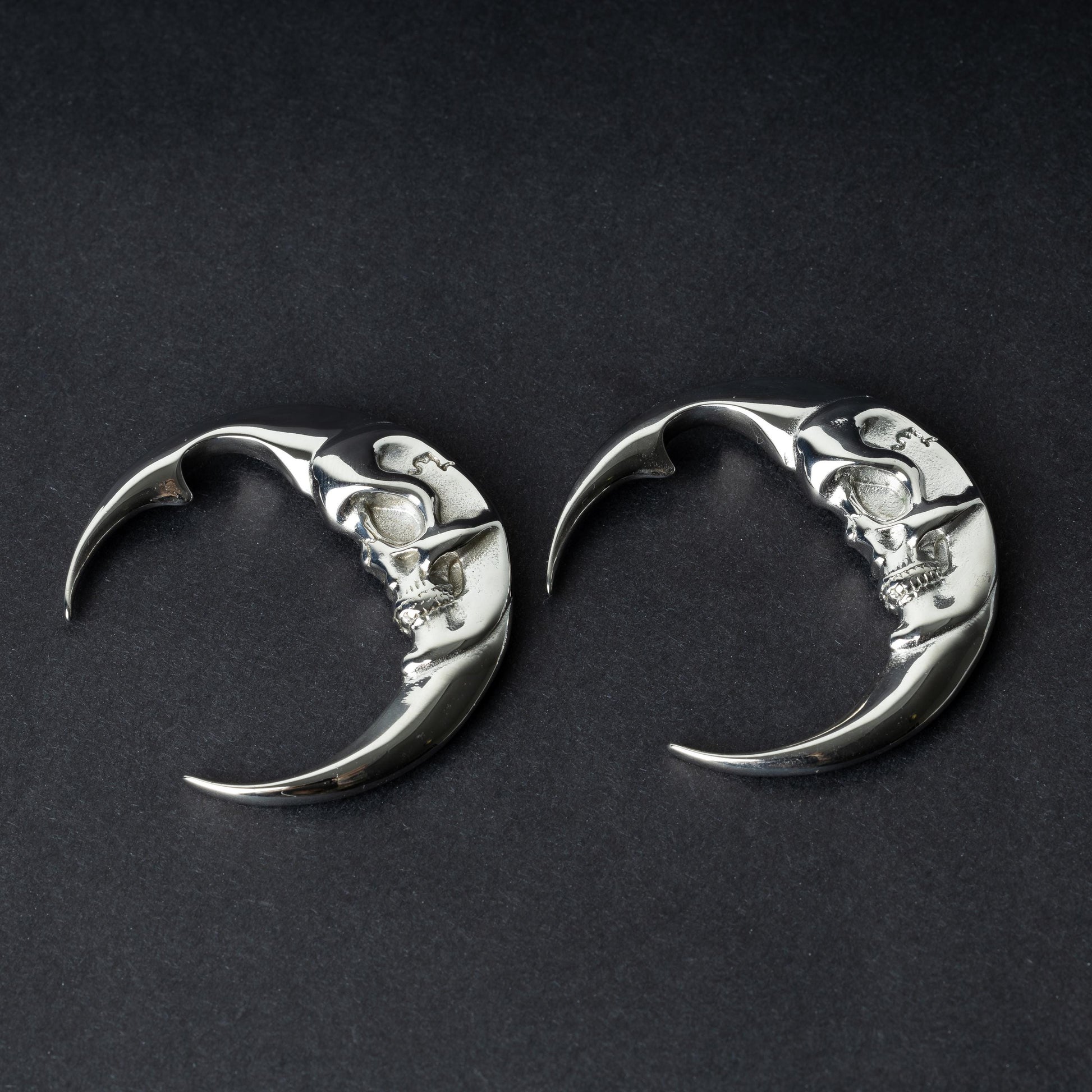 A pair of crescent moon-shaped ear weights in polished silver with detailed moon face designs, displayed on a dark background.

