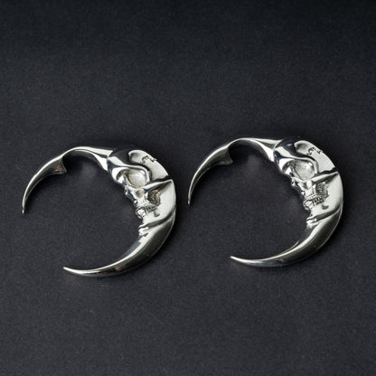 A pair of crescent moon-shaped ear weights in polished silver with detailed moon face designs, displayed on a dark background.

