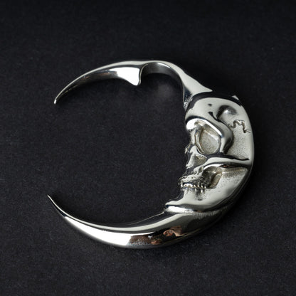 A pair of crescent moon-shaped ear weights in polished silver with detailed moon face designs, displayed on a dark background.

