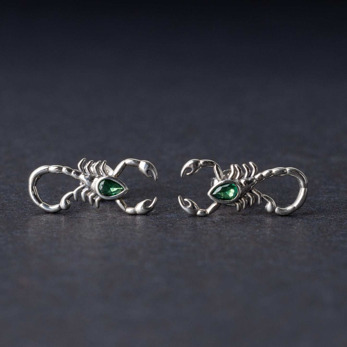 A pair of 925 sterling silver scorpion-shaped earrings with green gemstone accents as eyes, displayed on a dark background.