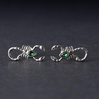 A pair of 925 sterling silver scorpion-shaped earrings with green gemstone accents as eyes, displayed on a dark background.