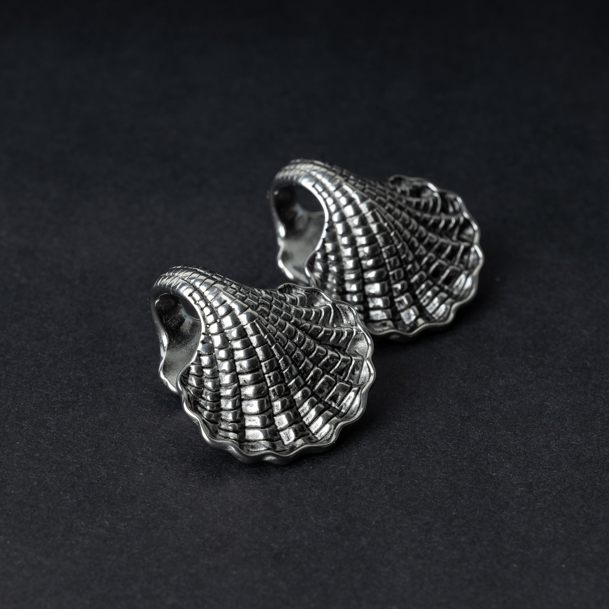 Two seashell-shaped ear weights in polished silver, with detailed textures and a bold design, displayed on a dark background.
