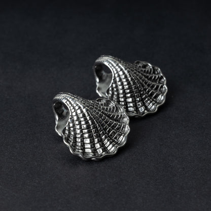 Two seashell-shaped ear weights in polished silver, with detailed textures and a bold design, displayed on a dark background.

