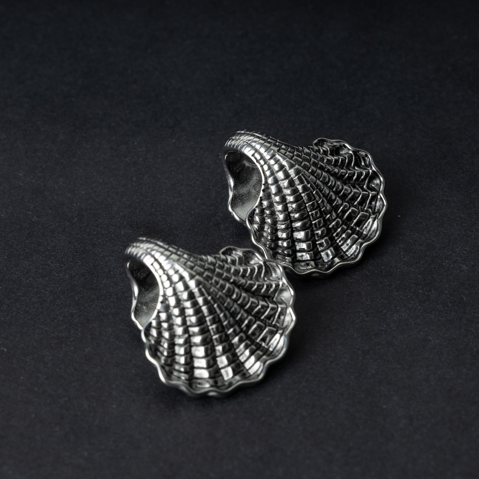 Two seashell-shaped ear weights in polished silver, with detailed textures and a bold design, displayed on a dark background.
