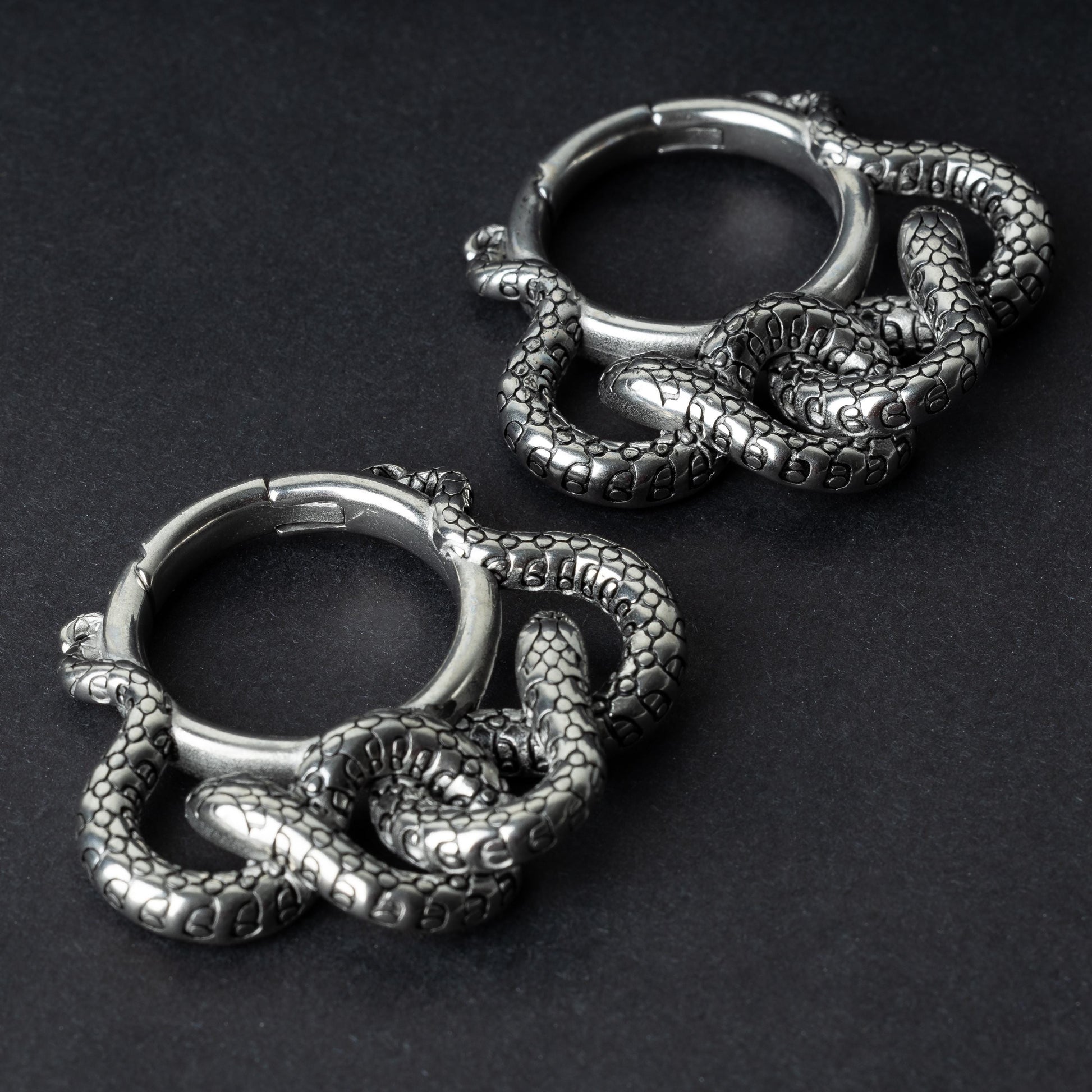 Two snake ear weights in polished silver, featuring detailed scales and a seamless hinge closure, displayed on a dark background.

