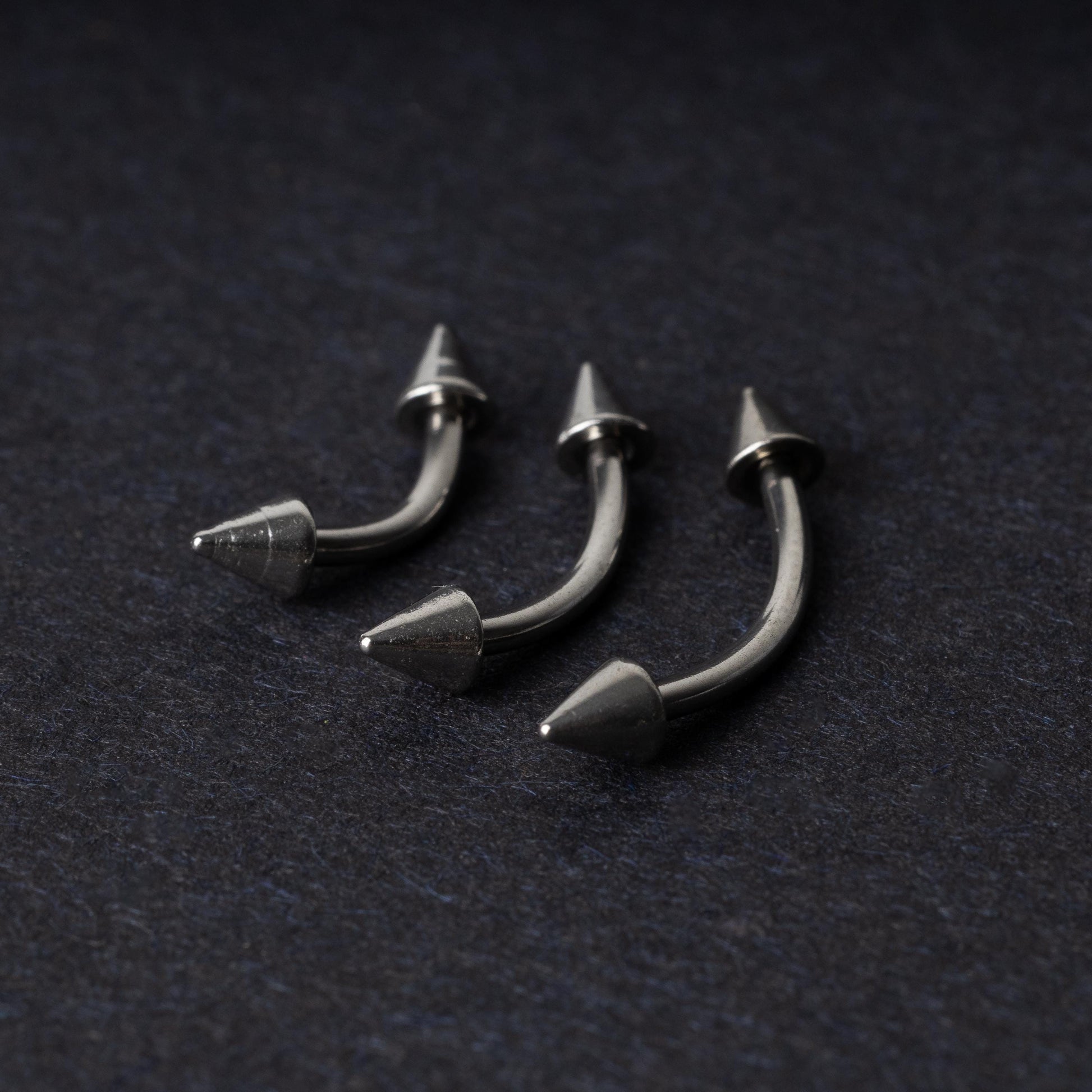 Three stainless steel curved barbells with cone-shaped ends, displayed on a dark background.
