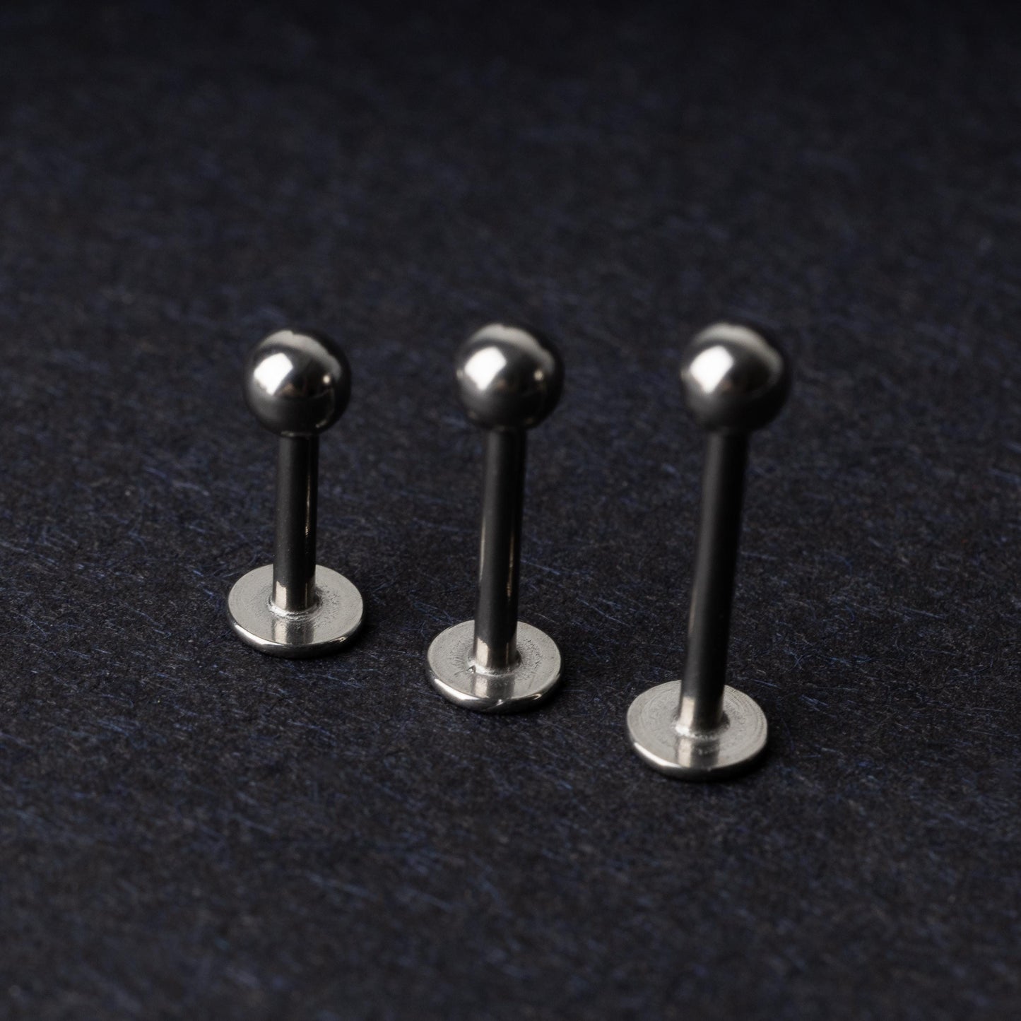 Three silver stainless steel labret studs with flat backs and polished ball ends, displayed on a dark textured background.