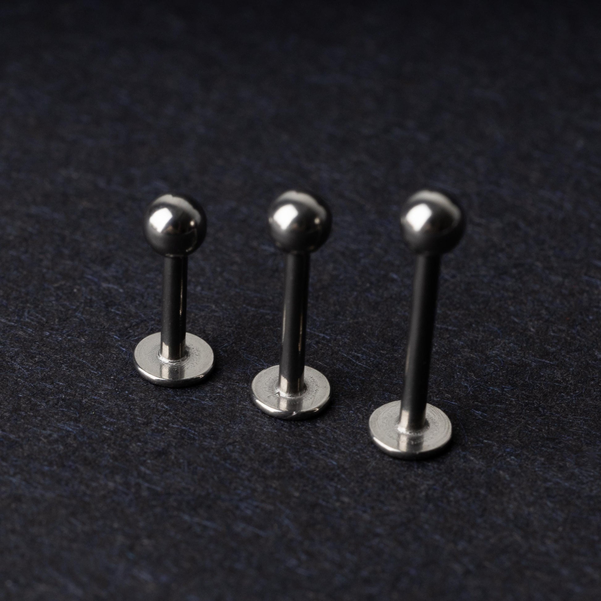 Three silver stainless steel labret studs with flat backs and polished ball ends, displayed on a dark textured background.