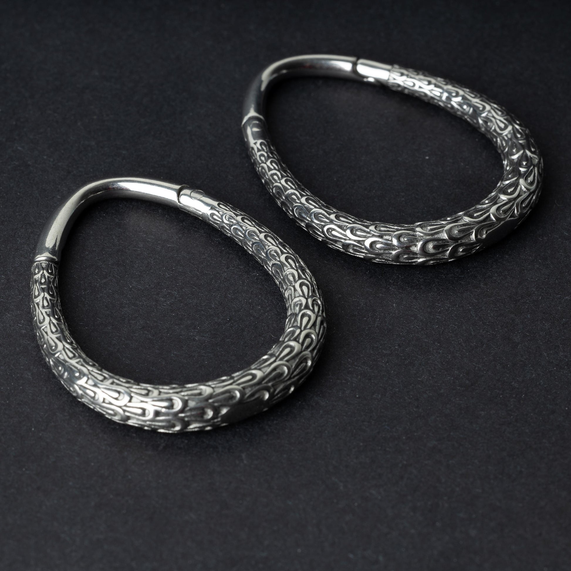 A pair of silver teardrop ear weights featuring a detailed dragon-scale texture and a seamless hinge closure, displayed on a dark background.

