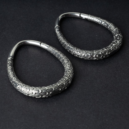 A pair of silver teardrop ear weights featuring a detailed dragon-scale texture and a seamless hinge closure, displayed on a dark background.

