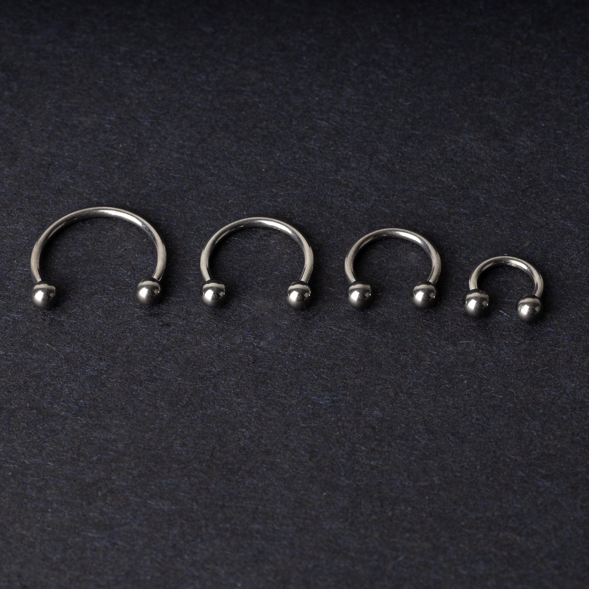 Four titanium horseshoe hoops with ball ends, arranged from largest to smallest on a dark background.