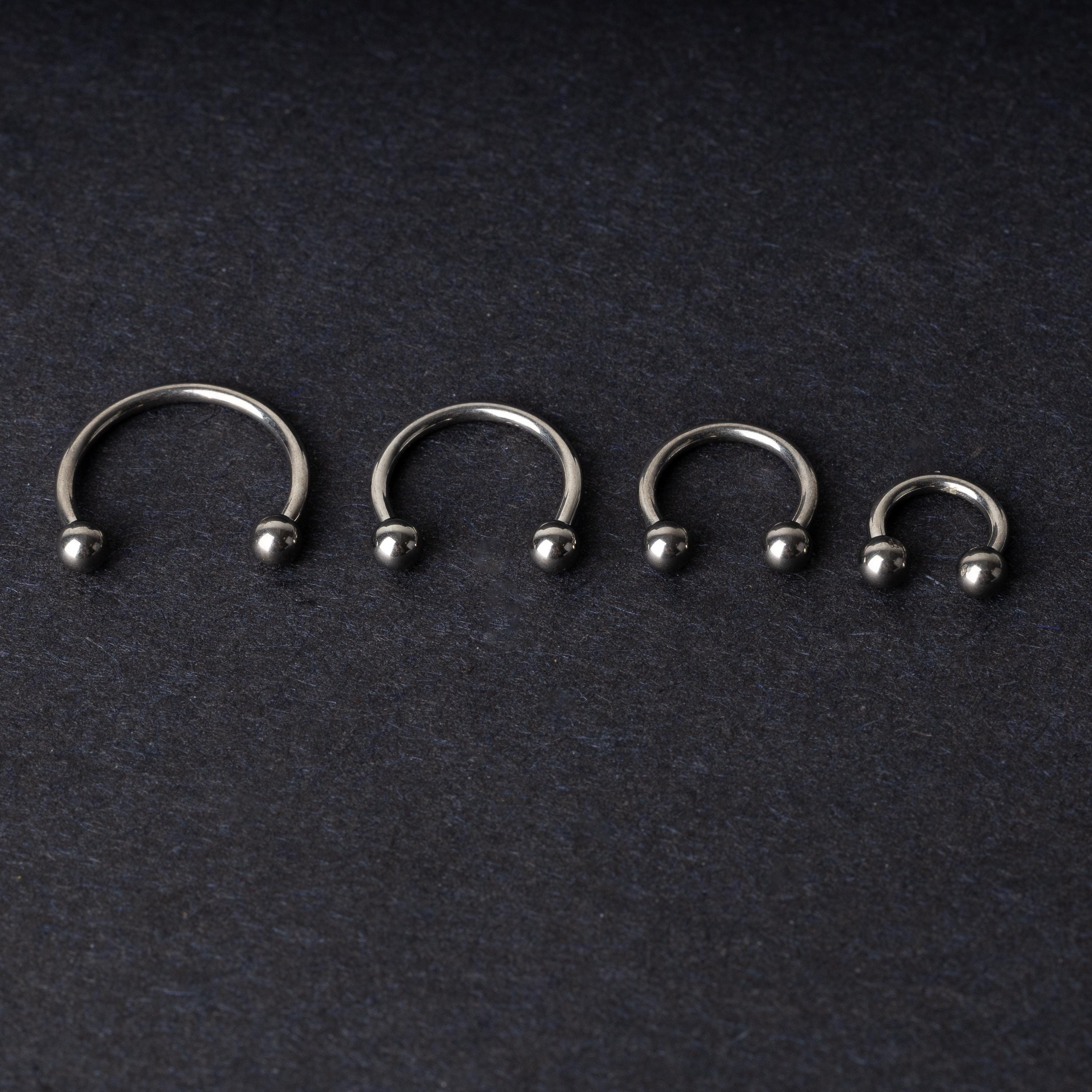Four titanium horseshoe hoops with ball ends, arranged from largest to smallest on a dark background.