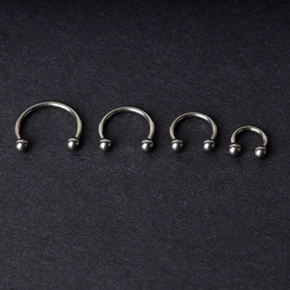 Four titanium horseshoe hoops with ball ends, arranged from largest to smallest on a dark background.
