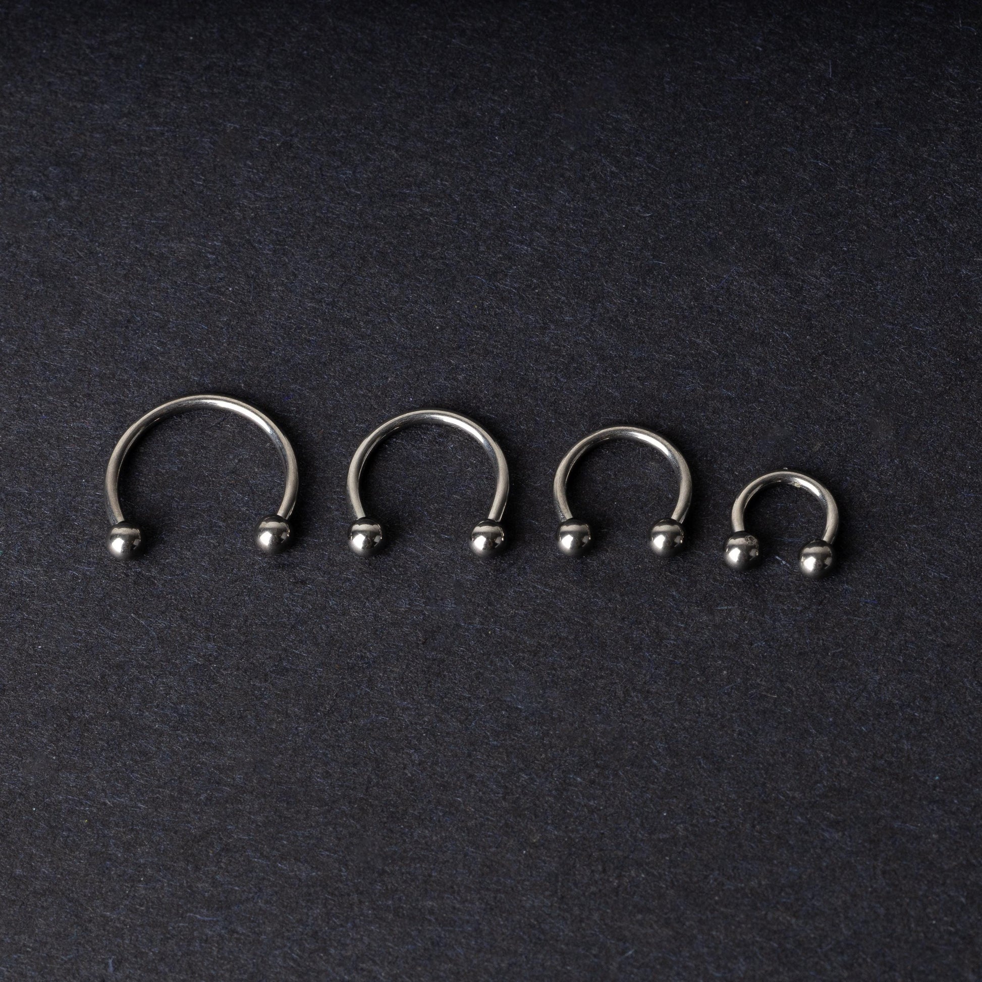 Four titanium horseshoe hoops with ball ends, arranged from largest to smallest on a dark background.