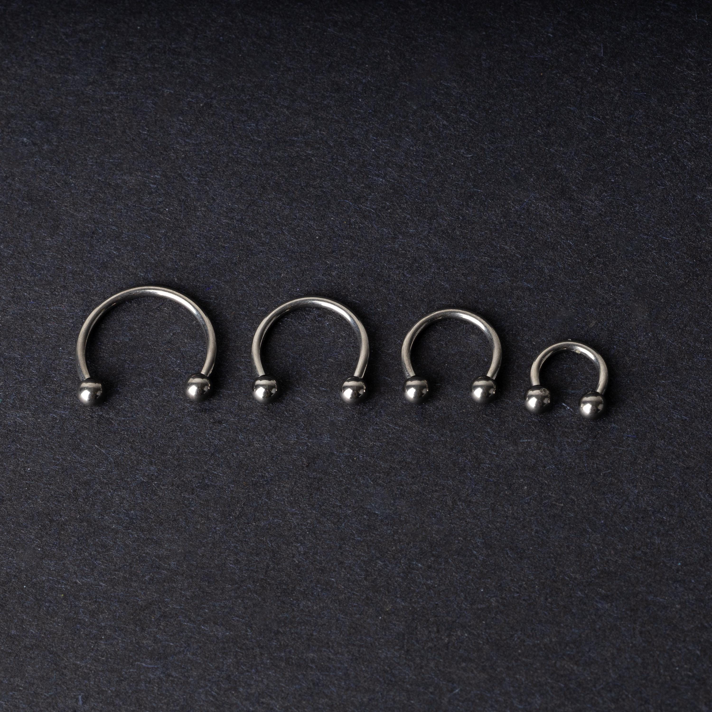 Four titanium horseshoe hoops with ball ends, arranged from largest to smallest on a dark background.