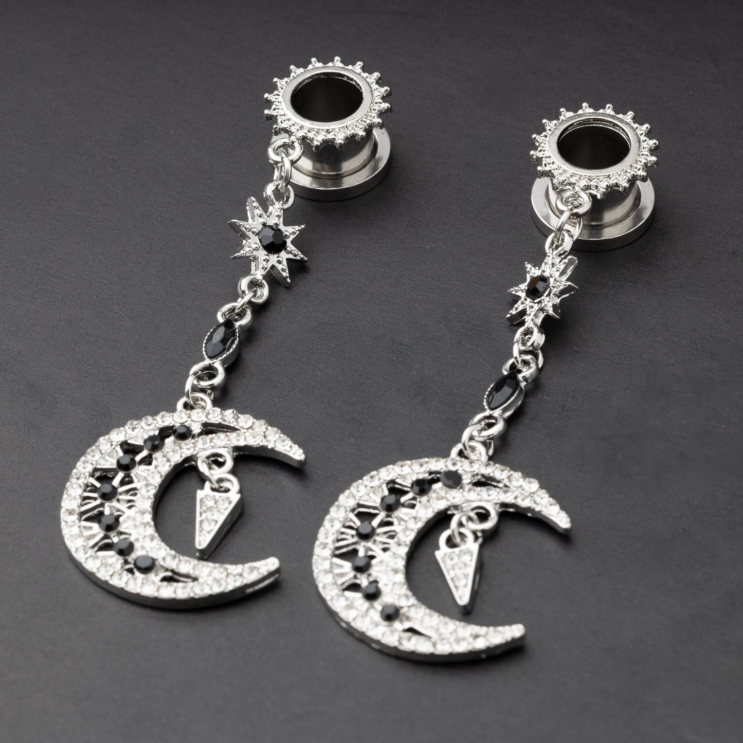 A pair of silver ear hangers for stretched ears with dangling crescent moon and star designs, adorned with black and clear rhinestones, displayed on a dark background.