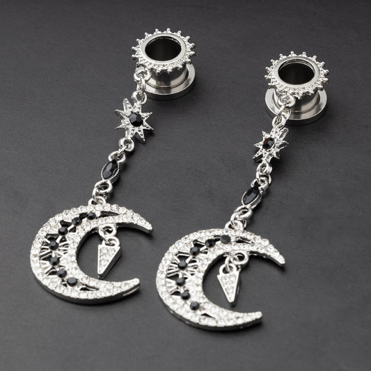A pair of silver ear hangers for stretched ears with dangling crescent moon and star designs, adorned with black and clear rhinestones, displayed on a dark background.