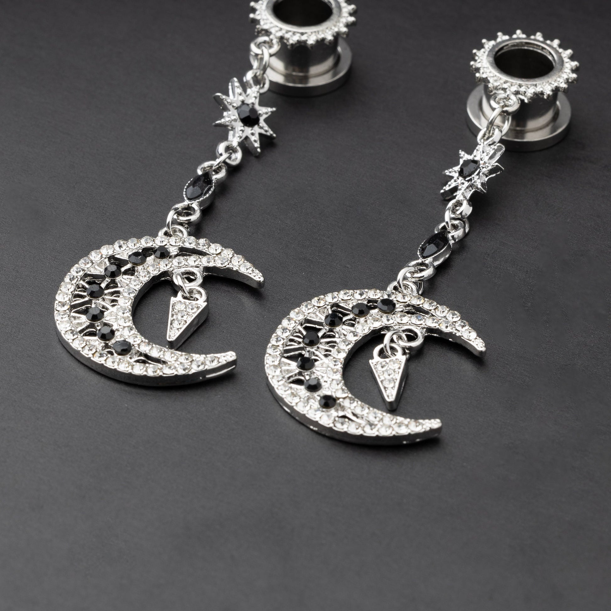A pair of silver ear hangers for stretched ears with dangling crescent moon and star designs, adorned with black and clear rhinestones, displayed on a dark background.