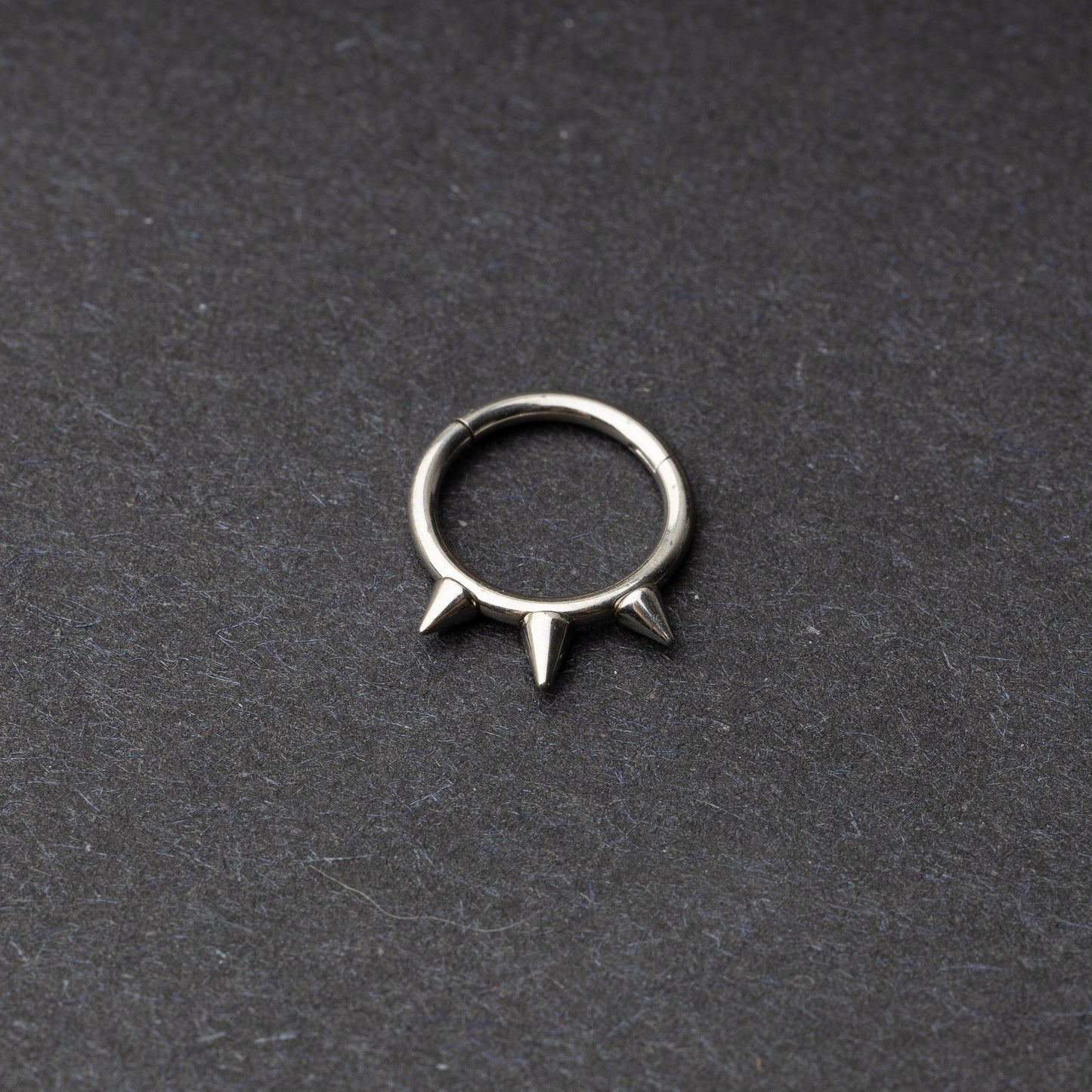 Silver-tone stainless steel clicker ring with three spikes, displayed on a dark textured background.