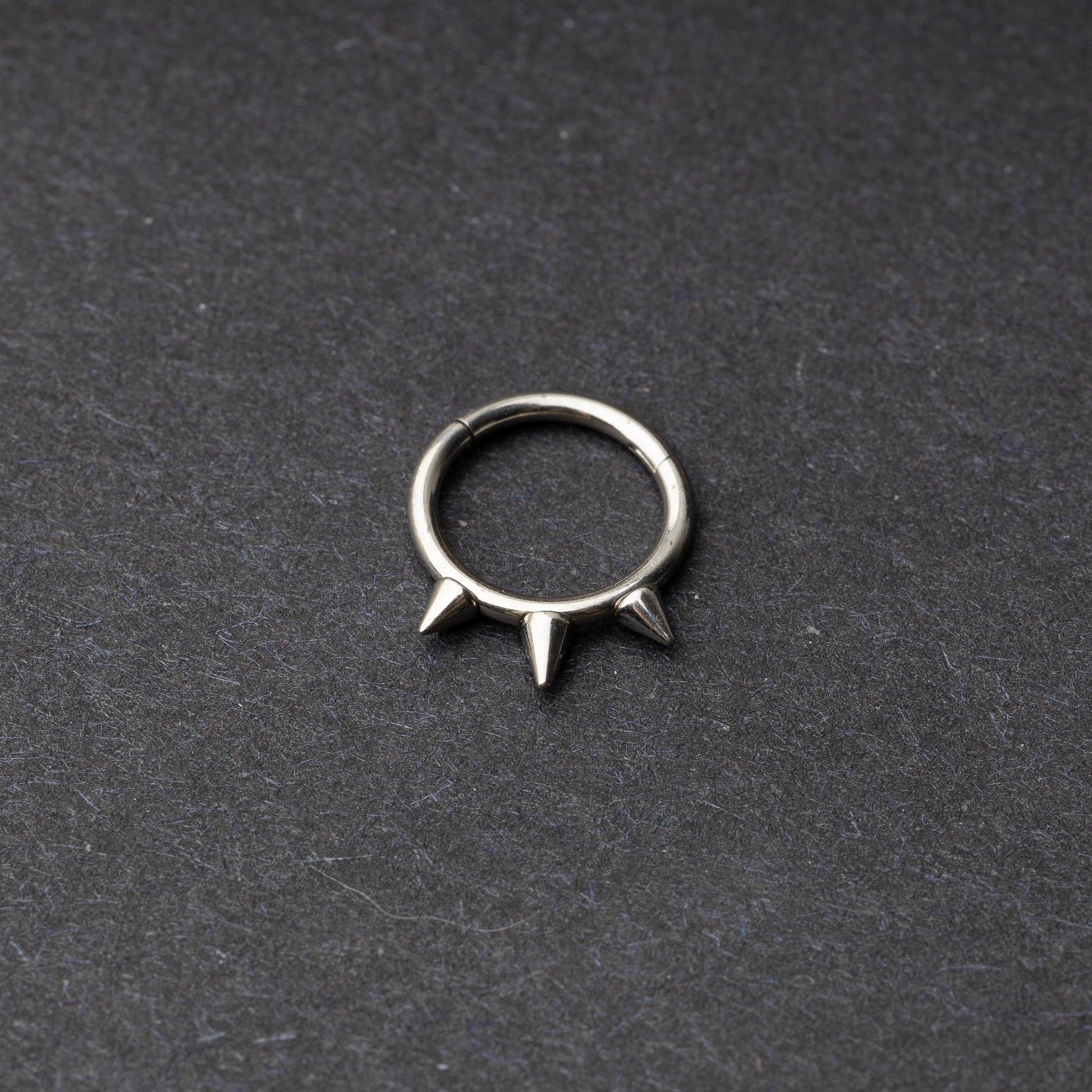 Silver-tone stainless steel clicker ring with three spikes, displayed on a dark textured background.