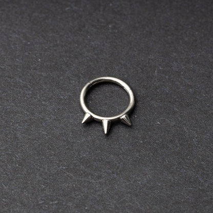 Silver-tone stainless steel clicker ring with three spikes, displayed on a dark textured background.