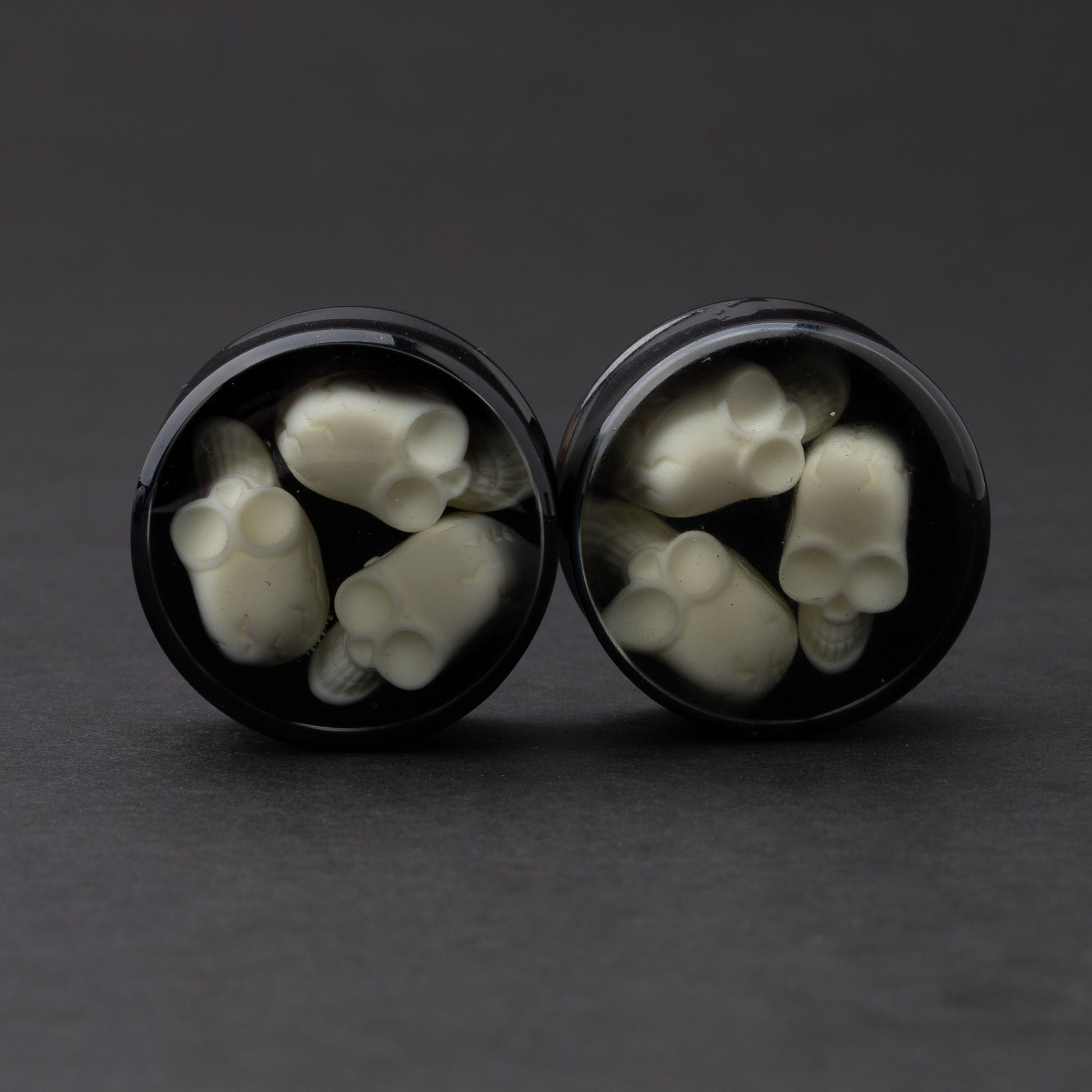 A pair of black double-flared plugs with glow-in-the-dark skulls encased in a polished finish, displayed on a dark background.