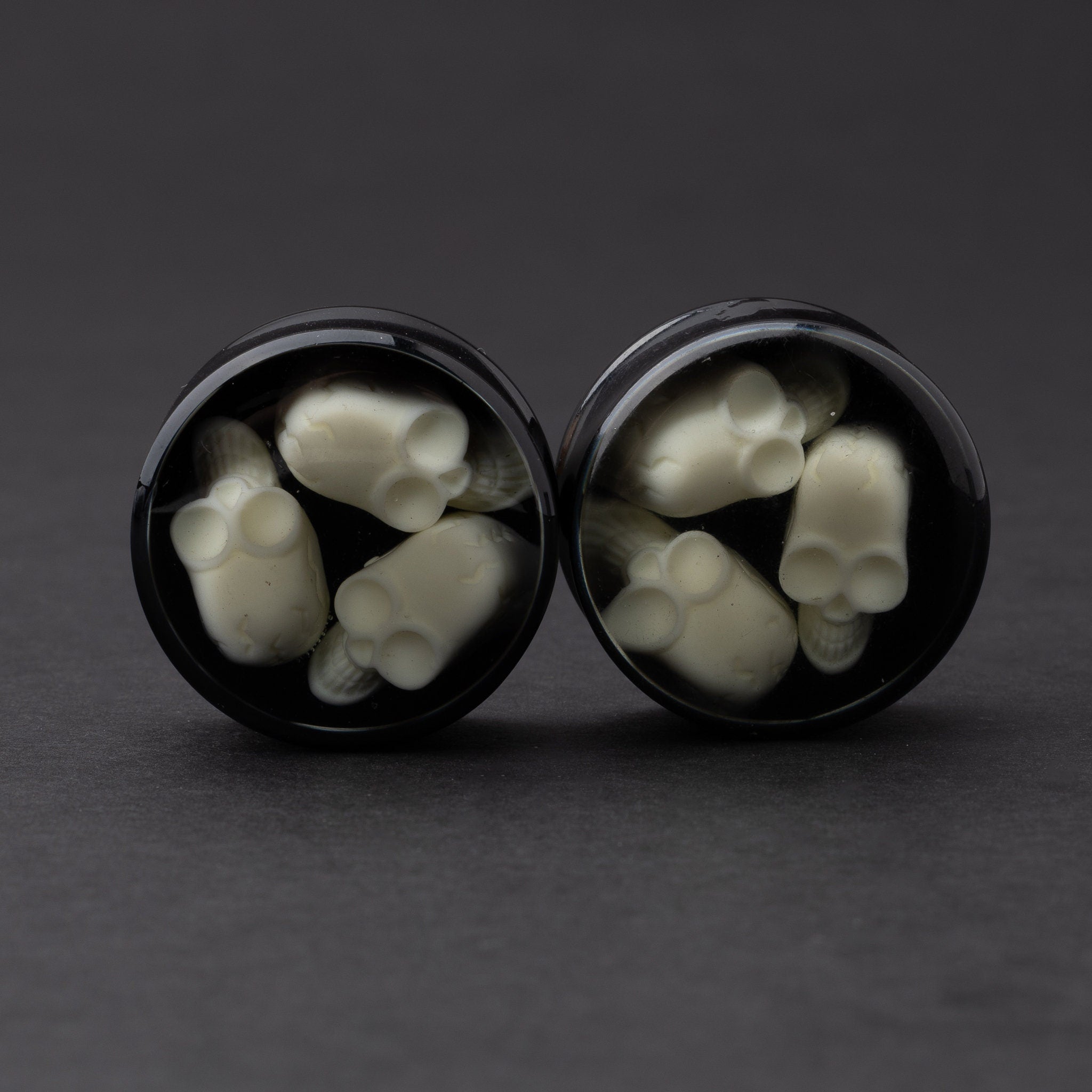 A pair of black double-flared plugs with glow-in-the-dark skulls encased in a polished finish, displayed on a dark background.