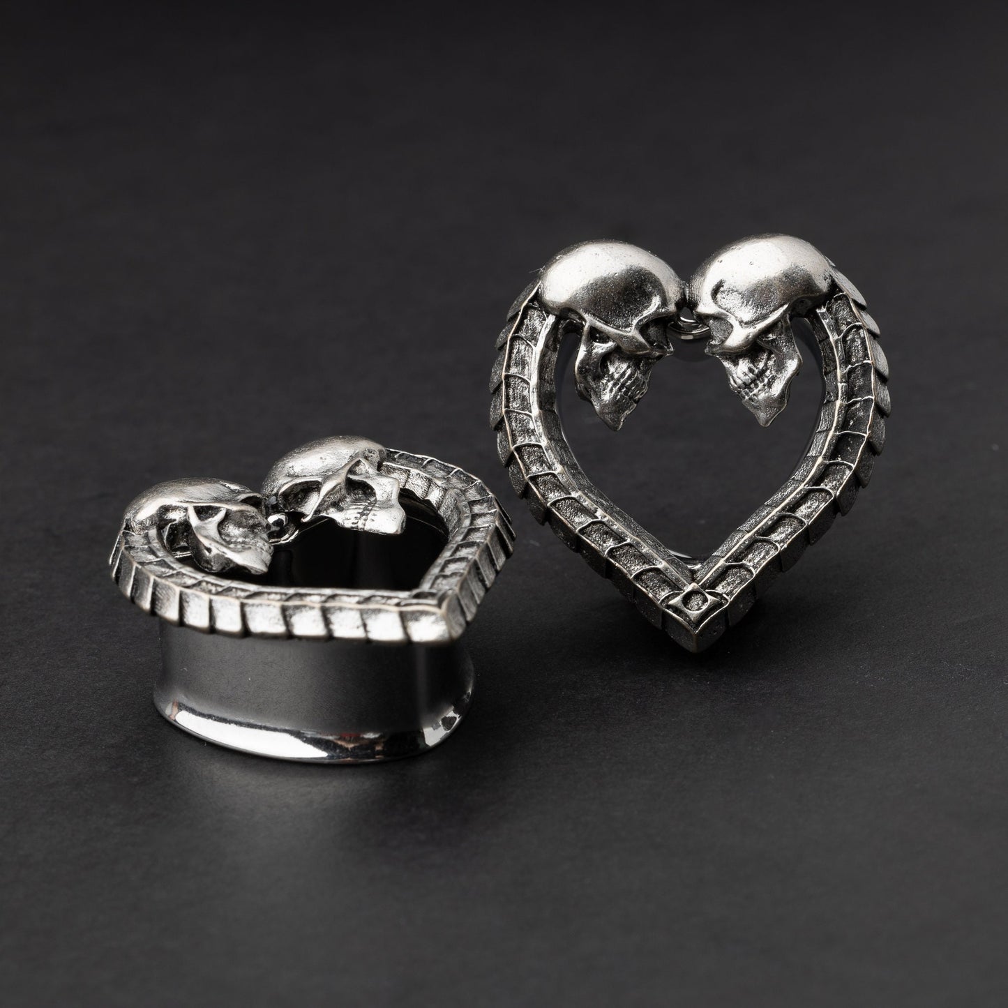 Heart-shaped tunnel plugs with intricate skull details and textured edges, displayed on a dark background.